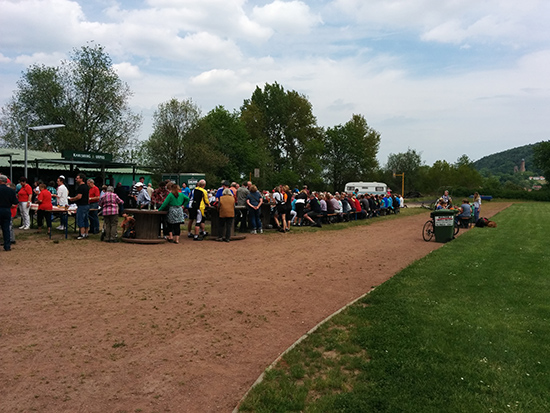 Viele Menschen sitzen auf Bierzeltgarnieturen vor einem Zelt in der Sonne, vorne wird Essen ausgegeben, im Hintergrund sieht man die Kirkeler Burg