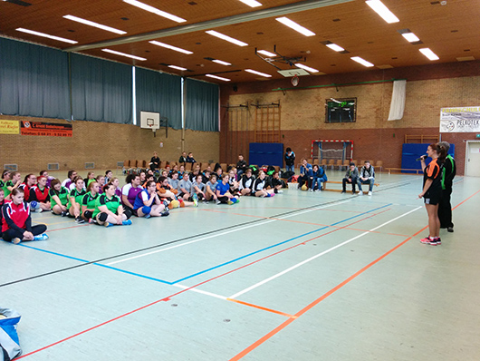 Alle Mannschaften sitzen auf dem Boden der Halle und vor ihnen steht die Turnierleiterin, um sie zu begrüßen.